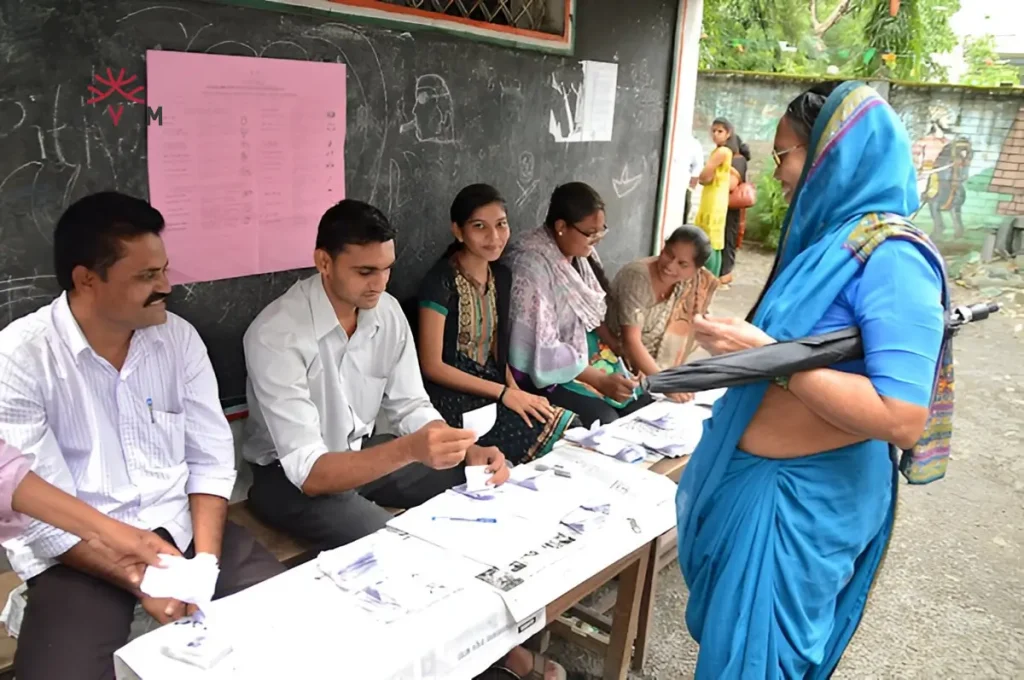 A woman wearing a sari stands with a group, representing the collective benefits of enrolling in the MGNREGA Scheme.
