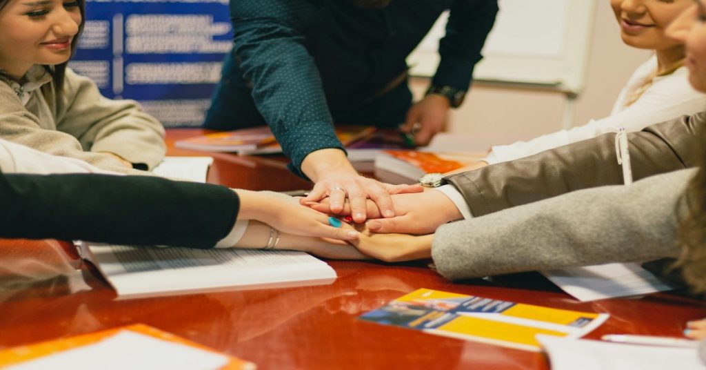 Individuals of various backgrounds holding hands around a table, representing collaboration in CSR sponsorships for NGOs.