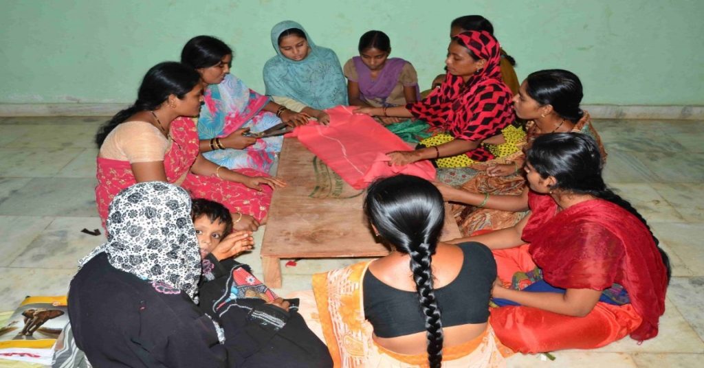 Women in saris gather around a table with fabric, learning new skill under MGNREGA scheme.