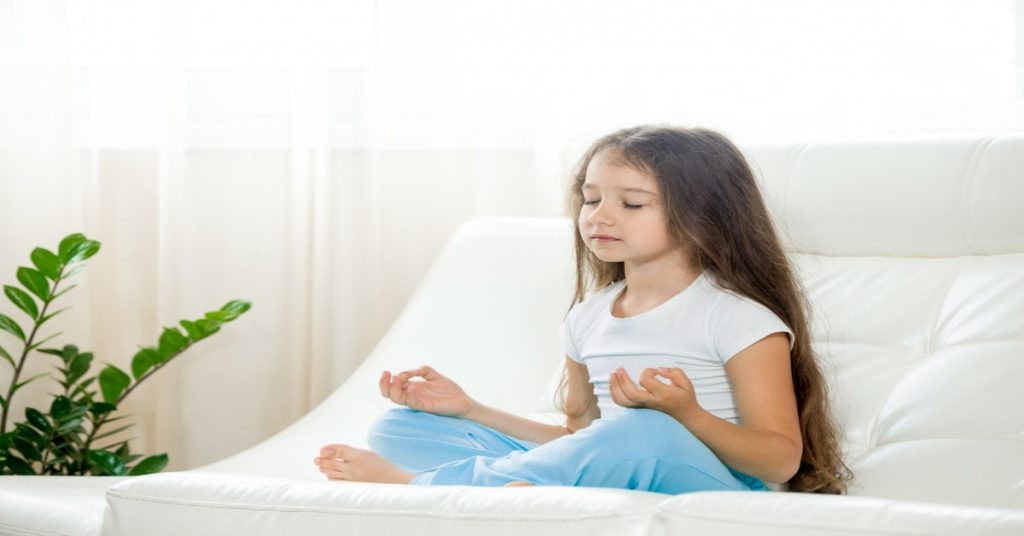 A little girl practicing yoga on a couch, embodying emotional intelligence through self-regulation and calm-down routines.