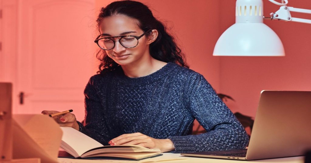 A woman in glasses is seated at a desk with a laptop and a book, learning online skills for content writing and copywriting.