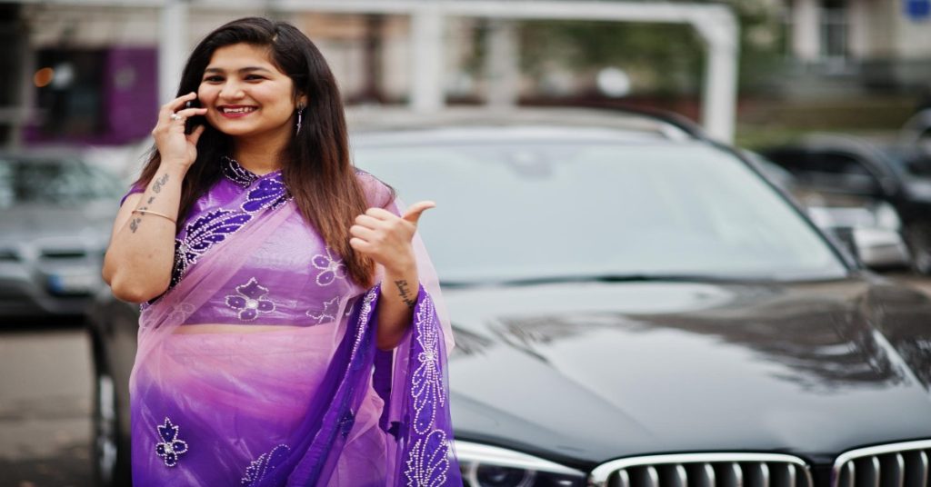 A woman in a purple sari on a phone call with her BMW car behind, emphasizing how online skills contribute to women's financial independence.